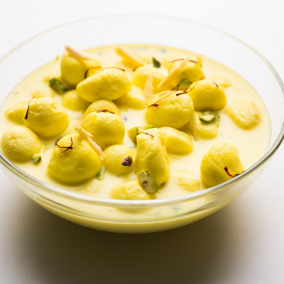 A glass bowl filled with yellow Rasmalai, garnished with saffron and pistachios in a creamy sauce.
