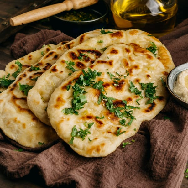 Freshly baked Indian naan bread, garnished with cilantro, on a brown cloth with oil and dip.
