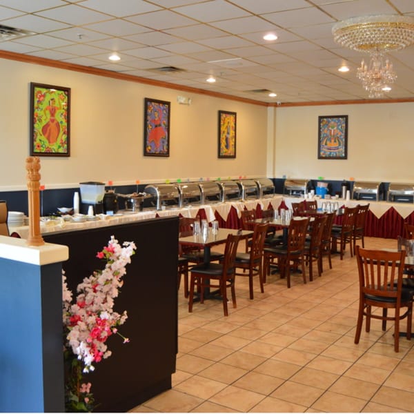 An Indian restaurant dining room with a buffet line, tables, chairs, and colorful artwork on the walls.