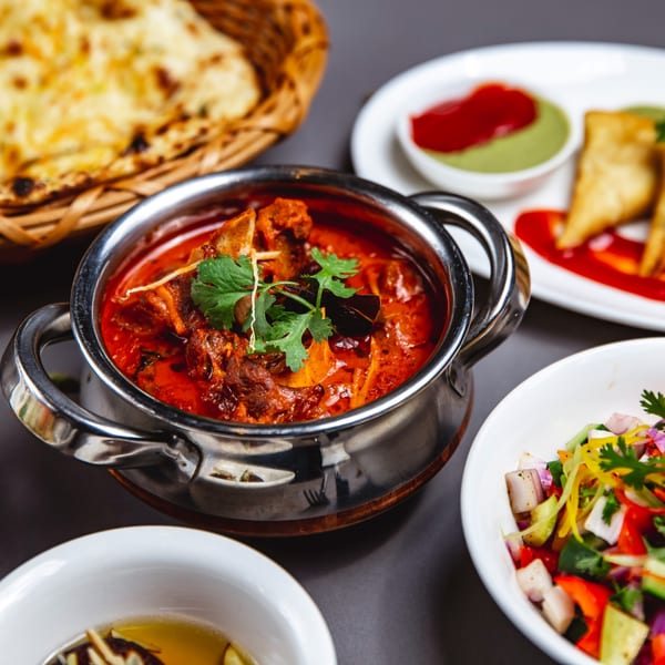 Indian curry in a metal pot with naan bread, samosas, and a fresh salad.