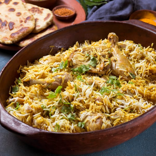 Chicken biryani with cilantro in a clay pot, with naan bread and spices in the background.